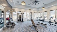 Located at 2720 Bristlecone Trail in Raleigh, NC, this fitness center offers a thoughtfully designed and generously sized exercise space complemented by a connecting hallway. The main exercise room is defined by soaring ceilings, large windows lining multiple walls, and an abundance of natural light that creates an open, energizing atmosphere with pleasant outdoor views of the surrounding landscape.

The flooring throughout the exercise area features a patterned, multi-tone carpet tile that adds texture and visual interest while providing durability underfoot. Stylish gold pendant lights and a large ceiling fan hang overhead, enhancing both the aesthetics and comfort of the space.

Equipment is comprehensive and well-organized. The cardio zone includes multiple treadmills, stationary bikes with integrated digital screens, elliptical machines, and a row of cycling bikes positioned along the windowed wall. A dedicated free weights area features a full rack of dumbbells alongside adjustable benches and floor space for stretching or functional training. A cable resistance machine and a squat/power rack round out the strength training offerings.

One wall is lined with large mirrors, amplifying the sense of space and supporting form-focused training. A wall-mounted television is positioned for easy viewing, and a small media/utility station adds everyday convenience. The adjacent hallway, finished with light hardwood-style flooring, provides a clean and polished transition into the fitness area.