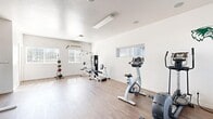 Located at 650 South Wolverine Way in Orem, Utah, this fitness center offers a clean, functional workout environment housed within a single open-plan space. The room features durable wood-style flooring throughout, crisp white walls, and a smooth ceiling with flush-mounted lighting that keeps the area bright and welcoming. Multiple windows fitted with horizontal blinds allow natural light to fill the space, enhancing the open, airy feel. A wall-mounted mini-split HVAC unit ensures year-round climate comfort during workouts. The facility is well-appointed with a diverse range of equipment, including a treadmill, upright stationary bike, elliptical trainer, and a multi-station cable and weight machine. A medicine ball rack with a variety of weighted balls supports functional training, while a dedicated dumbbell rack provides free weight options. A pull-up and dip station rounds out the strength training offerings. The open floor plan allows ample room for stretching, floor exercises, or movement between stations. A decorative wolf emblem on the wall adds a subtle community identity to the space. The layout and variety of equipment make this fitness center a practical and accessible resource for a broad range of workout routines.