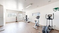 Located at 650 South Wolverine Way in Orem, Utah, this fitness center offers a clean, functional workout environment housed within a single open-plan space. The room features durable wood-style flooring throughout, crisp white walls, and a smooth ceiling with flush-mounted lighting that keeps the area bright and welcoming. Multiple windows fitted with horizontal blinds allow natural light to fill the space, enhancing the open, airy feel. A wall-mounted mini-split HVAC unit ensures year-round climate comfort during workouts. The facility is well-appointed with a diverse range of equipment, including a treadmill, upright stationary bike, elliptical trainer, and a multi-station cable and weight machine. A medicine ball rack with a variety of weighted balls supports functional training, while a dedicated dumbbell rack provides free weight options. A pull-up and dip station rounds out the strength training offerings. The open floor plan allows ample room for stretching, floor exercises, or movement between stations. A decorative wolf emblem on the wall adds a subtle community identity to the space. The layout and variety of equipment make this fitness center a practical and accessible resource for a broad range of workout routines.