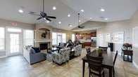 This single-floor space boasts an expansive open-concept layout that seamlessly integrates a living area, dining space, and full kitchen into one cohesive environment. The vaulted ceilings soar overhead, creating an airy, grand atmosphere enhanced by recessed lighting and ceiling fans that ensure both style and comfort throughout.

The living area is anchored by a classic white-mantled fireplace framed by decorative columns, providing a warm and inviting focal point. Comfortable seating, including a grey sofa and patterned accent chairs, is arranged to encourage conversation and relaxation. Large windows along the walls flood the space with natural light, lending a bright and welcoming ambiance.

Adjacent to the living area, the kitchen features rich wood cabinetry, stainless steel appliances including a full-size refrigerator, pendant lighting above the counters, and ample workspace — making it well-suited for both everyday use and entertaining. The dining area nearby accommodates a dark wood table and chairs, offering a defined space for meals without sacrificing the open feel of the room.

The flooring throughout features a light-toned tile laid in an elegant geometric pattern, tying together the various zones of the space with a cohesive and polished aesthetic. Overall, the space combines functional design with refined finishes for a comfortable and versatile environment.