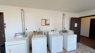 This single-floor laundry facility is designed for shared use, providing a practical and efficiently organized space for handling laundry needs. The room features multiple top-loading washing machines and dryers arranged in a row along the walls, each unit equipped with coin-operated mechanisms for convenient, self-service use. The exhaust venting for the dryers is routed through stainless steel duct pipes that extend vertically along the walls, reflecting a straightforward and functional mechanical setup.

The interior walls are finished in a clean white, contributing to a bright and utilitarian atmosphere. The flooring consists of tile in a neutral tone, offering durability and ease of maintenance. The ceiling features a textured finish typical of shared utility spaces. A cork bulletin board mounted on the wall displays informational notices and guidelines for users of the facility.

The overall layout is open and uncluttered, allowing for easy movement between machines. The space prioritizes practicality, with its design focused on providing residents or users with a reliable and accessible laundry solution. The clean, no-frills aesthetic ensures the facility remains easy to maintain and straightforward to navigate.