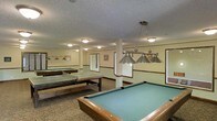This single-floor recreational space is thoughtfully designed to accommodate a variety of indoor games and leisure activities. The centerpiece of the room is a full-sized pool table finished in rich wood tones, positioned beneath a classic suspended pendant light fixture with brushed nickel shades that provide focused, functional illumination. Additional pool tables are visible throughout the space, along with a ping-pong table, making this an ideal setting for group entertainment. The room features neutral, light-toned walls accented with a decorative border trim that adds a subtle architectural detail. Flush-mount ceiling fixtures supplement the pendant lighting, ensuring the entire area is evenly and warmly lit. The textured ceiling and carpeted flooring contribute to a comfortable, casual ambiance. Structural columns are integrated naturally into the layout, defining zones within the open floor plan without interrupting the flow of the space. Windows along the walls allow for natural light to complement the interior lighting. Overall, this game room offers a spacious, inviting environment well-suited for recreation and social interaction.