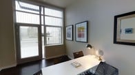 This three-floor space presents a modern, minimalist aesthetic characterized by clean architectural lines and thoughtful design details. The visible work or dining area exemplifies the overall tone of the space, featuring large floor-to-ceiling windows fitted with vertical blinds that invite abundant natural light while offering views of the surrounding exterior environment. The dark hardwood flooring grounds the room and contrasts effectively with the light gray walls, creating a balanced, contemporary palette.

A sleek white table serves as the central furnishing, accompanied by dark wood chairs that add warmth and contrast. Decorative touches, including a brass desk lamp, a small globe, and two framed abstract artworks mounted on the wall, contribute to an intellectually curated atmosphere. The ceiling height in this area is notably generous, enhancing the sense of openness.

The space benefits from direct access to the outdoors through a glass-paneled door adjacent to the large window, further connecting the interior to the natural environment outside. Across its three floors, the property likely offers versatile and well-lit spaces that maintain this consistent modern sensibility, combining functional design with refined aesthetic choices.