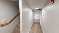 This two-floor residence offers a well-organized and functional layout with consistent, modern finishes throughout. The kitchen serves as a central highlight, featuring crisp white shaker-style cabinetry, dark quartz countertops, a peninsula with an undermount sink, and a full suite of stainless steel appliances including a refrigerator, electric range, and microwave. The open layout connects the kitchen to a bright living area, enhanced by a ceiling fan and a window that allows natural light to fill the space. Light hardwood-style flooring runs throughout the main living areas, lending warmth and continuity to the design. A carpeted staircase with a wood handrail connects the two floors, accompanied by a clean, well-lit hallway with flush-mounted ceiling lights and a vertical blind-covered window at the end. The bathrooms are finished in a neutral palette, featuring tub-shower combinations with curved curtain rods, hardwood-style flooring, and vanities with stone-look countertops and large mirrors. White paneled doors and bright white walls contribute to an overall clean and airy ambiance, making the space feel open and easy to maintain.