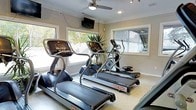 This single-story fitness room is designed to support an effective cardio workout in a bright, welcoming environment. The space is outfitted with multiple Star Trac treadmills and an elliptical trainer, providing residents or users with quality cardio equipment. Large windows along the walls allow generous natural light to fill the room while offering views of surrounding greenery, creating an airy and motivating atmosphere. A ceiling fan with an integrated light fixture helps maintain comfortable airflow during workouts. A wall-mounted flat-screen television is positioned for easy viewing from the cardio machines, adding entertainment to exercise sessions. Crown molding along the ceiling lends a polished architectural detail to the room, elevating it beyond a purely utilitarian space. A large mirror on one wall visually expands the room and allows users to monitor their form. Decorative indoor plants add a touch of natural life to the interior. The neutral, light-toned walls and flooring contribute to a clean, modern aesthetic throughout. Overall, the space balances functional fitness amenities with thoughtful design details.