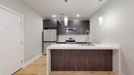 This single-floor space showcases a contemporary design aesthetic throughout, with light hardwood flooring and a neutral color palette creating a cohesive, modern atmosphere. The open-concept kitchen serves as a central highlight, featuring flat-front dark wood cabinetry that extends to the ceiling for ample storage. A large waterfall-edge quartz island with an undermount sink and a sleek pull-down faucet anchors the space, while matching marble-look quartz countertops and a coordinating tile backsplash run along the perimeter. Stainless steel appliances, including a French door refrigerator, electric range, and built-in microwave, complete the culinary space. Cylindrical pendant lights hang above the island, complementing the recessed lighting throughout. The bathroom is finished with large-format white tile flooring, a marble-look walk-in shower enclosed by a frameless sliding glass door, and a warm wood vanity. A dedicated walk-in closet features built-in shelving and double hanging rods. The in-unit laundry area accommodates a stacked washer and dryer with adjacent shelving. A bedroom offers natural light through a large window and continues the hardwood flooring. A wall-mounted electric fireplace adds warmth to the living area.