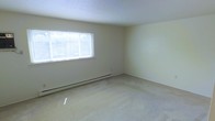 This single-floor space presents a clean, minimalist interior defined by its simple and functional design. The main room is generously proportioned, offering an open and airy feel that makes the most of its footprint. Neutral, cream-toned walls create a calm and versatile backdrop, while wall-to-wall beige carpeting adds warmth underfoot and contributes to the overall understated aesthetic.

A wide, horizontal window fitted with white blinds serves as the room's primary source of natural light, allowing ample daylight to filter in and illuminate the space. Glimpses of greenery visible through the window suggest a pleasant outdoor setting. A baseboard heating unit runs along the exterior wall beneath the window, providing an efficient and unobtrusive heating solution.

The textured ceiling adds a subtle visual element consistent with a practical, well-maintained interior. The room's layout is open and flexible, lending itself well to a variety of furniture arrangements and uses. Overall, the space conveys a sense of quiet simplicity, with its light-filled atmosphere and neutral palette offering a welcoming and adaptable environment.