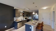 Located at 201 S Elliot Rd in Chapel Hill, NC, this single-floor residence showcases a thoughtfully designed interior with modern finishes throughout. The open-concept kitchen serves as the centerpiece of the home, featuring rich dark wood cabinetry that provides ample storage alongside light-toned walls, creating a striking visual contrast. A granite-topped island with seating for multiple guests doubles as a dining and entertaining space, illuminated by elegant pendant lighting that adds warmth to the area. The kitchen is fully equipped with a full suite of stainless steel appliances, including a French door refrigerator with ice and water dispenser, a built-in microwave, a dishwasher, and a range. A track lighting system runs along the ceiling, providing adjustable task lighting across the cooking area. Light wood-style flooring runs throughout, contributing to a clean, contemporary aesthetic. Open shelving integrated into the cabinetry design offers an opportunity to display décor and everyday essentials. The overall design balances functionality with modern style, making the space well-suited for both daily living and entertaining guests.
