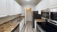 This single-floor unit offers a practical and well-appointed layout designed for comfortable everyday living. The main living area is finished with dark hardwood-style flooring that runs throughout, complemented by neutral gray walls and a textured ceiling. A sliding glass door fitted with vertical blinds allows natural light to fill the space and provides access to the exterior. A programmable thermostat is conveniently mounted on the wall for climate control.

The galley-style kitchen is efficiently designed, featuring white cabinetry on both sides, dark granite-look countertops, a stainless steel double sink, and a full suite of appliances including a refrigerator, electric range, microwave, and dishwasher. Light wood-tone flooring in the kitchen provides a clean contrast to the cabinetry, and a doorway at the end of the kitchen leads to the laundry area, which includes an in-unit top-loading washer with overhead wire shelving.

The bedroom is carpeted for warmth and comfort, with a ceiling fan providing overhead lighting and air circulation. A window with horizontal blinds admits natural light, creating a bright and restful atmosphere. The bathroom includes a clean, full-size soaking tub and shower combination. Throughout the unit, consistent finishes and thoughtful details contribute to a cohesive and move-in-ready living environment.
