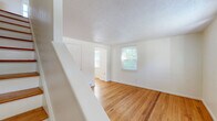 Located at 6A Weis Rd in Albany, NY, this 2-bedroom, 1-bathroom home spans three floors, offering a well-organized and functional layout throughout. The main level features a bright living room with warm hardwood flooring, white walls, and a front entry door with a transom window that allows natural light to fill the space. A staircase leads to the upper levels, maintaining a clean, open feel. The kitchen is equipped with white cabinetry, dark laminate countertops, a gas range, and a double basin sink beneath a window overlooking the outdoors. An adjoining dining room completes the main living area. Upstairs, two bedrooms continue the hardwood flooring theme. The primary bedroom includes a closet and benefits from multiple windows that bring in generous natural light and views of the surrounding greenery. The second bedroom is compact yet functional. The full bathroom showcases classic tilework with a black-and-navy accent border, a checkerboard mosaic tile floor, a pedestal-style vanity, and a tub-shower combination with a window above. The basement level provides unfinished utility space, housing the water heater and offering additional storage potential. Textured ceilings are present throughout the upper floors, adding a traditional character to the home.