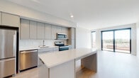 This contemporary single-floor residence is designed with a clean, modern aesthetic that emphasizes open living and functional elegance. The heart of the home is an open-concept kitchen and living area, seamlessly connected to create a bright, airy atmosphere. The kitchen is outfitted with sleek, flat-front grey cabinetry extending to the ceiling for ample storage, a crisp white quartz countertop, and a full suite of stainless steel appliances including a refrigerator, electric range, microwave, and dishwasher. A central island provides additional workspace and serves as a natural gathering point. Light wood-tone flooring flows throughout the space, complementing the neutral white walls and creating a warm, cohesive feel. The living area benefits from generous natural light streaming through floor-to-ceiling sliding glass doors that open onto a private balcony, offering open city views. The bathroom features a clean, minimalist design with a soaking tub and shower combination, a wide vanity with a white basin sink, brushed gold fixtures, and a large mirror. The overall ambiance of this residence is one of understated sophistication, combining modern design sensibilities with practical, comfortable living.