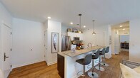 This single-floor residence offers a thoughtfully designed layout that balances modern style with everyday comfort. The open-concept kitchen is a standout feature, equipped with stainless steel appliances, white cabinetry, a mosaic tile backsplash, and a spacious peninsula with quartz-style countertops illuminated by striking pendant lights. A row of contemporary bar stools makes the peninsula ideal for casual dining or entertaining.

The living room exudes a warm, relaxed ambiance with recessed lighting, wood-look flooring, and a wall-mounted television above a decorative lattice-door sideboard. The adjacent dining area accommodates a full-sized table for six, accented by a large mirror and natural light from nearby windows with linen drapery.

The primary bedroom is generously sized, featuring plush carpeting, large windows that invite natural light, and ample space for a full bedroom suite including a dresser with geometric detailing. A second bedroom doubles as a functional workspace, with room for both a full bed and a wooden writing desk. Each bedroom benefits from natural light through well-positioned windows.

Two bathrooms serve the residence: one with a double-sink vanity featuring dark cabinetry and a wide mirror, and a second with a single-sink vanity, tile flooring, and a shower-tub combination. Both bathrooms are clean and well-finished, offering practical convenience throughout.