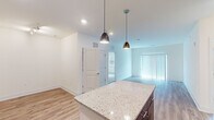 This single-floor residence offers a thoughtfully designed open-concept layout that seamlessly connects the kitchen, dining, and living areas. The kitchen serves as the centerpiece of the space, showcasing a large granite-topped island illuminated by sleek pendant lights, two-tone cabinetry with light wood upper cabinets and dark lower cabinets, and a full suite of stainless steel appliances including a range, microwave, dishwasher, and refrigerator. A crisp white tile backsplash and light hardwood-style flooring complete the kitchen's modern aesthetic. The living area extends naturally from the kitchen, bathed in natural light from a large sliding glass door fitted with vertical blinds. The hardwood-style flooring continues throughout this open zone, creating a cohesive and airy feel. The bedroom is a comfortable retreat featuring plush carpeting, a ceiling fan with light fixture, and a generously sized walk-in closet with built-in shelving. Natural light filters through a window with blinds, adding warmth to the room. The bathroom is clean and functional, featuring tile flooring, a granite-topped vanity with wood cabinetry, and a full soaking tub with shower combination. Throughout the space, neutral tones and recessed lighting contribute to a bright, modern, and welcoming atmosphere.