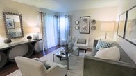 This single-story living space showcases a well-appointed living room designed with a refined, contemporary aesthetic. The room is anchored by a patterned area rug that defines the seating arrangement, which includes a grey upholstered sofa accented with teal geometric throw pillows, a pair of white nailhead-trim armchairs, and a sleek glass-and-metal coffee table. Dark hardwood flooring runs throughout, providing a warm contrast to the soft grey and white tones of the walls and furnishings. Natural light flows generously into the space through a large window dressed with layered curtains — sheer white panels paired with patterned drapery — offering both brightness and privacy. A sliding glass door adjacent to the window connects the interior to an outdoor area, further enhancing the sense of openness. The walls are tastefully decorated with a framed botanical print and an arrangement of decorative mirrors in varied geometric shapes. A dark console table near the entrance holds a table lamp and small accessories, adding functional elegance. A textured ceiling completes the space, giving it a classic residential character throughout.