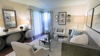 This single-story living space showcases a well-appointed living room designed with a refined, contemporary aesthetic. The room is anchored by a patterned area rug that defines the seating arrangement, which includes a grey upholstered sofa accented with teal geometric throw pillows, a pair of white nailhead-trim armchairs, and a sleek glass-and-metal coffee table. Dark hardwood flooring runs throughout, providing a warm contrast to the soft grey and white tones of the walls and furnishings. Natural light flows generously into the space through a large window dressed with layered curtains — sheer white panels paired with patterned drapery — offering both brightness and privacy. A sliding glass door adjacent to the window connects the interior to an outdoor area, further enhancing the sense of openness. The walls are tastefully decorated with a framed botanical print and an arrangement of decorative mirrors in varied geometric shapes. A dark console table near the entrance holds a table lamp and small accessories, adding functional elegance. A textured ceiling completes the space, giving it a classic residential character throughout.