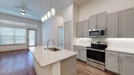 This well-appointed single-floor residence offers a thoughtfully designed open-concept layout that seamlessly connects the kitchen and living areas. The kitchen serves as the centerpiece of the space, showcasing a large island with a granite countertop and an undermount sink, illuminated by a cascading cluster of pendant lights. Grey shaker-style cabinetry lines the walls, complemented by a herringbone tile backsplash and a full suite of stainless steel appliances, including a range, over-the-range microwave, and refrigerator. Warm hardwood-style flooring flows consistently throughout the main living areas, grounding the bright, neutral color palette of the walls and trim. The living area benefits from generous natural light, courtesy of large windows and a glass-panel exterior door, while a ceiling fan adds both function and comfort. Elevated ceilings contribute to an airy, open ambiance across the main spaces. The bathroom continues the refined aesthetic with matching grey cabinetry, granite countertops, a framed mirror, and large-format tile accents near the tub surround. Built-in shelving near the entryway adds practical storage. Overall, the space presents a clean, contemporary design with consistent high-quality finishes throughout.