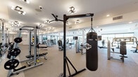 This expansive fitness center is designed to accommodate a comprehensive range of workout routines within a single, open floor plan. The space is bright and airy, benefiting from large windows along the perimeter that allow abundant natural light and offer views to the outdoors. Neutral-toned carpet flooring, crisp white walls, and a combination of recessed lighting, ceiling fans, and stylish multi-bulb fixtures create a clean, modern atmosphere throughout.

The cardio zone is well-appointed with multiple treadmills, elliptical trainers, a rowing machine, and a stair climber, many positioned to face mounted flat-screen televisions. A dedicated free weights area features a full rack of dumbbells alongside adjustable benches and large wall mirrors that enhance the sense of space. TKO-branded strength machines and a cable system provide ample options for resistance training, while a freestanding TKO heavy bag and boxing station adds a dynamic training element.

A separate stretching and functional training area includes floor-to-ceiling mirrors, motivational wall graphics, and space for floor exercises. A tastefully designed amenity nook features textured tile accent walls, a warm wood bench with coat hooks, a beverage refrigerator, and a wine rack — offering a refined post-workout retreat. The overall design is polished and functional, catering to fitness enthusiasts at every level.