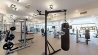This expansive fitness center is designed to accommodate a comprehensive range of workout routines within a single, open floor plan. The space is bright and airy, benefiting from large windows along the perimeter that allow abundant natural light and offer views to the outdoors. Neutral-toned carpet flooring, crisp white walls, and a combination of recessed lighting, ceiling fans, and stylish multi-bulb fixtures create a clean, modern atmosphere throughout.

The cardio zone is well-appointed with multiple treadmills, elliptical trainers, a rowing machine, and a stair climber, many positioned to face mounted flat-screen televisions. A dedicated free weights area features a full rack of dumbbells alongside adjustable benches and large wall mirrors that enhance the sense of space. TKO-branded strength machines and a cable system provide ample options for resistance training, while a freestanding TKO heavy bag and boxing station adds a dynamic training element.

A separate stretching and functional training area includes floor-to-ceiling mirrors, motivational wall graphics, and space for floor exercises. A tastefully designed amenity nook features textured tile accent walls, a warm wood bench with coat hooks, a beverage refrigerator, and a wine rack — offering a refined post-workout retreat. The overall design is polished and functional, catering to fitness enthusiasts at every level.