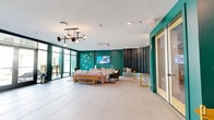 This vibrant amenity lounge offers a well-appointed, open-concept layout that seamlessly blends a social lounge area with a fully equipped kitchen. The space is defined by its bold emerald green accent walls, which create a striking visual contrast against the bright white tile flooring and white ceiling with recessed lighting. A sculptural chandelier with globe bulbs anchors the lounge area, which features a plush pink sofa, velvet green armchairs, a slatted wood bench, and decorative butterfly wall art that add personality and warmth.

A standout architectural feature is the curved, floor-to-ceiling upholstered wall panel finished in charcoal gray, which forms a dramatic semi-circular seating nook with built-in cushioned benches. Adjacent to this is a wall-mounted flat-screen TV and a wood-toned credenza, making the space equally suited for relaxed gatherings or casual presentations.

The kitchen area is equally impressive, centered around a large curved island with a richly veined dark granite countertop and a fluted wood base, surrounded by stylish pink barstools. Light wood cabinetry, open shelving with artful décor, white subway tile backsplash, and stainless steel appliances complete the kitchen. Expansive floor-to-ceiling glass walls and sliding doors flood the space with natural light and provide direct access to an outdoor terrace.