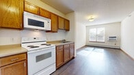 This single-floor unit offers a practical and straightforward layout designed for comfortable everyday living. The kitchen is equipped with warm oak cabinetry extending both above and below the countertops, providing ample storage space. White appliances — including a coil-burner electric range, over-the-range microwave, refrigerator, and dishwasher — are neatly arranged along the kitchen wall. A single-basin sink and light-toned countertops complete the functional workspace. The kitchen opens directly into the living area, which features wood-look hard-surface flooring that extends throughout the main spaces, allowing natural light from a window to brighten the room. A window air conditioning unit and baseboard heating system ensure year-round climate control. The bedroom is carpeted for added comfort and includes a window with blinds that invites natural light, along with a closet for storage. The bathroom is designed with accessibility in mind, featuring a spacious walk-in shower with grab bars and a handheld showerhead, complemented by oak vanity cabinetry and tile flooring. A coat closet with wire shelving near the entry adds additional storage convenience throughout the unit.