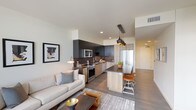 Located at 1777 Wewatta Street in the heart of Denver, Colorado, this single-floor residence offers a well-designed open-concept layout that seamlessly connects the living, dining, and kitchen areas. The living room is anchored by floor-to-ceiling windows and a glass door that opens to a private balcony, framing expansive views of the Denver skyline. Natural light fills the space throughout the day, complementing the wood-style flooring and neutral color palette.

The kitchen is outfitted with a striking two-tone cabinetry scheme, pairing dark upper cabinets with light wood lower units. A textured tile backsplash, stainless steel appliances—including a built-in microwave, electric range, and French door refrigerator—and a central island with bar seating complete the space. Pendant lighting adds a refined finishing touch.

The bedroom continues the modern aesthetic with large windows offering prominent city views, providing ample natural light. The bathroom features light wood-toned cabinetry, an illuminated mirror, and a full tub-shower combination with clean white tile surroundings. Throughout the residence, the design prioritizes clean lines, functional layouts, and contemporary finishes suited to urban living.