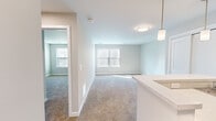 This 1-bedroom, 1-bathroom unit offers a thoughtfully designed single-floor layout that maximizes every square foot. The open-concept living and dining area is finished with plush wall-to-wall carpeting and soft, neutral-toned walls that create a bright, welcoming atmosphere. Natural light flows in through multiple windows fitted with blinds, complemented by contemporary flush-mount and pendant lighting fixtures.

The galley-style kitchen is equipped with dark espresso cabinetry, light countertops, a stainless steel sink, and a full suite of black appliances including a range, microwave, refrigerator, and dishwasher. Light hardwood-style flooring anchors the kitchen, contrasting cleanly with the cabinetry. A peninsula counter with pendant lighting above opens toward the living space, adding functionality and visual flow.

The bedroom offers a comfortable footprint with soft carpeting, neutral walls, and direct access to a spacious walk-in closet outfitted with wire shelving and hanging rods along multiple walls — providing ample storage. The full bathroom features hexagonal tile flooring, a dark-toned vanity with a white countertop and rectangular mirror, and a walk-in shower with chrome fixtures. Clean lines and a neutral palette give the bathroom a polished, modern feel throughout.