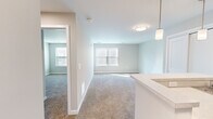 This 1-bedroom, 1-bathroom unit offers a thoughtfully designed single-floor layout that maximizes every square foot. The open-concept living and dining area is finished with plush wall-to-wall carpeting and soft, neutral-toned walls that create a bright, welcoming atmosphere. Natural light flows in through multiple windows fitted with blinds, complemented by contemporary flush-mount and pendant lighting fixtures.

The galley-style kitchen is equipped with dark espresso cabinetry, light countertops, a stainless steel sink, and a full suite of black appliances including a range, microwave, refrigerator, and dishwasher. Light hardwood-style flooring anchors the kitchen, contrasting cleanly with the cabinetry. A peninsula counter with pendant lighting above opens toward the living space, adding functionality and visual flow.

The bedroom offers a comfortable footprint with soft carpeting, neutral walls, and direct access to a spacious walk-in closet outfitted with wire shelving and hanging rods along multiple walls — providing ample storage. The full bathroom features hexagonal tile flooring, a dark-toned vanity with a white countertop and rectangular mirror, and a walk-in shower with chrome fixtures. Clean lines and a neutral palette give the bathroom a polished, modern feel throughout.