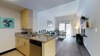 This single-floor residence offers a thoughtfully designed open-concept layout that seamlessly integrates the kitchen and living area into one cohesive and functional space. The kitchen is appointed with light-toned cabinetry, granite countertops, a stainless steel sink, and a built-in dishwasher, all centered around a peninsula that doubles as a dining and prep area. The space flows naturally into the living room, where wide-plank hardwood-style flooring extends throughout, creating a warm and cohesive aesthetic underfoot. The living area is bright and inviting, anchored by a large window dressed in sheer white curtains that allows an abundance of natural light to fill the room while framing views of the surrounding neighborhood. The white walls and high ceilings enhance the sense of openness and airiness throughout the space. The overall ambiance is clean, modern, and welcoming, with a neutral color palette that provides a versatile backdrop for a variety of interior styles. This well-appointed residence balances contemporary design with everyday functionality, making it an ideal space for both relaxed living and casual entertaining.
