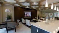 This single-floor space presents a thoughtfully designed lounge or common area that balances modern aesthetics with functional comfort. The most striking architectural feature is the ceiling, which showcases an elaborate geometric pattern of layered, circular coffers with integrated recessed lighting. This distinctive design element adds considerable visual depth and sophistication to the room.

The main lounge area centers around a large wall-mounted flat-screen television set against a rich, dark wood-paneled accent wall, which serves as a bold focal point. Comfortable cream and white upholstered armchairs and seating clusters are arranged to encourage conversation and relaxation, accented by dark wood side tables and burgundy throw pillows.

Light-toned large-format tile flooring with a subtle herringbone pattern runs throughout, contributing to the bright, open feel of the space. A partial kitchen or bar counter with a light-colored quartz surface is visible in the foreground, suggesting an adjacent catering or refreshment area. In the background, a dining or café-style area with bar seating and pendant lighting provides additional functionality. Overall, the space conveys an upscale, hospitality-inspired ambiance suited for social gatherings.