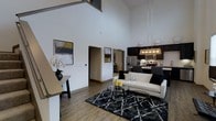 This thoughtfully designed two-story residence offers two bedrooms and two bathrooms across a spacious, well-appointed floor plan. The heart of the home is an impressive open-concept living and kitchen area distinguished by soaring double-height ceilings that create an airy, expansive atmosphere. Large white walls provide a clean, modern canvas accented by contemporary artwork, while wood-look flooring runs throughout the main level, adding warmth and continuity.

The living area is generously sized, anchored by a sleek glass coffee table and a neutral sofa, complemented by a bold patterned area rug that adds texture and contrast. Natural light flows freely through the open layout, enhancing the sense of space.

The kitchen is equipped with dark cabinetry, stainless steel appliances including a refrigerator and microwave, and a light-colored countertop that provides striking contrast to the rich cabinetry. A stylish pendant light fixture illuminates the kitchen workspace, adding a decorative focal point.

A lofted upper level overlooks the main living space below, taking full advantage of the dramatic ceiling height. The two-floor layout offers a clear separation between living and private spaces, making this residence both functional and visually dynamic — ideal for those seeking modern, open living.