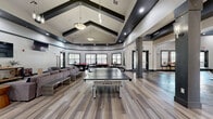 Located at 201 Inman St in Denton, TX, this single-floor community clubhouse offers a generously sized, amenity-rich environment designed for both recreation and productivity. The space is characterized by soaring ceilings with bold dark beam accents, luxury vinyl plank flooring throughout, and a cohesive modern aesthetic anchored by a neutral white and charcoal color palette.

The welcoming entryway features a dramatic hallway with star-shaped wall décor, a sleek glass-top display table, and a statement chandelier overhead, setting an inviting tone. From there, the space opens into a sprawling game and lounge area equipped with pool tables, a ping-pong table, a shuffleboard table, and a large sectional sofa arrangement facing wall-mounted flat-screen TVs — ideal for social gatherings.

A dedicated casual dining and lounge zone includes high-top tables and bar seating beneath a distinctive curved dark ceiling, complemented by a large round game table and vending amenities nearby. The study and conference area provides multiple long work tables with vibrant teal chairs, dark wood cabinetry, and an abundance of natural light through a stacked grid of windows. A leasing center is also accessible within the facility. Throughout, pendant lighting, recessed fixtures, and strategic accent lighting enhance the polished, contemporary ambiance of this well-appointed community space.