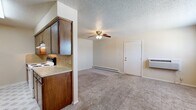 This single-floor, 2-bedroom, 1-bathroom residence offers a practical and straightforward layout designed for comfortable everyday living. The main living area features wall-to-wall carpeting, a ceiling fan with light fixture, baseboard heating, and a wall-mounted HVAC unit, ensuring year-round climate comfort. A window with blinds brings in natural light, creating a bright and open feel. The galley-style kitchen is efficiently designed with rich, warm-toned wooden cabinetry lining both sides, laminate countertops with a tile backsplash, and patterned vinyl flooring. It is equipped with a white refrigerator, dishwasher, and electric range, along with ample upper and lower cabinet storage and a built-in sink. The primary bedroom is a well-proportioned space with soft carpeting and direct access to a dedicated closet featuring dual hanging rods and shelving, providing generous storage. The second bedroom also offers carpeted flooring and neutral walls. The full bathroom includes a wooden vanity with a single sink and countertop, a large mirror, and a separate water closet housing the tub-shower combination. Textured ceilings are present throughout, adding a consistent design element to this move-in-ready residence.