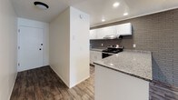 This single-floor unit offers a well-designed layout that combines modern finishes with distinctive architectural details throughout. The open-concept living and kitchen area is immediately welcoming, featuring durable wood-look flooring that flows seamlessly across the main spaces. A prominent brick accent wall serves as a standout design element in the living room, adding texture and character, while vertical blinds along large windows allow ample natural light to fill the space. The kitchen is thoughtfully appointed with white shaker-style cabinetry, granite countertops, a gray brick tile backsplash, a double-basin sink, and stainless steel appliances including an electric range. A peninsula counter provides additional workspace and creates a natural division between the kitchen and living area. The bedroom features soft carpeting and its own brick accent wall, echoing the design language found throughout the unit. The bathroom is equally refined, showcasing granite countertops, white cabinetry, a large vanity mirror, wood-look flooring, a separate soaking tub, and a tiled walk-in shower, all set against a textured brick wall. Textured ceilings and recessed lighting further contribute to the cohesive, updated aesthetic of this well-appointed space.