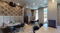 This thoughtfully designed single-floor communal space blends contemporary style with functional versatility across several distinct seating areas. The layout is anchored by a welcoming lounge zone bathed in natural light from expansive floor-to-ceiling windows dressed in striped linen drapes, offering views of the surrounding outdoor environment. Sleek swivel chairs arranged around petite tables create an inviting area for casual conversation or individual use.

A dedicated work area lines one wall, featuring a continuous granite-topped counter accompanied by cognac leather cantilever chairs and a striking diamond-pattern accent wall adorned with a row of warm gold sconce lighting — an eye-catching focal point that adds sophistication to the space.

Semi-private dining nooks provide an intimate contrast, each framed by tall upholstered banquette seating and bold patterned wallcovering in rich earth tones. Pendant drum lighting overhead and round tables paired with charcoal side chairs complete each nook's cozy, restaurant-inspired aesthetic. Textural room dividers in dark wood tones define zones without fully enclosing them.

Throughout, patterned carpeting and light wood-look flooring add visual depth, while curated abstract artwork punctuates the walls. The overall ambiance is polished yet comfortable, well-suited for socializing, focused work, or casual dining.