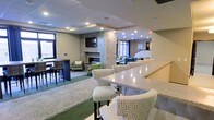 This single-floor communal space is designed to accommodate a variety of social settings within a polished, contemporary environment. A striking stone fireplace serves as the room's focal point, flanked by a mosaic tile accent that adds texture and visual interest. The fireplace area is accompanied by comfortable lounge seating in neutral tones accented with green throw pillows, creating a warm and inviting gathering spot. A wall-mounted flat-screen television above the fireplace enhances the entertainment value of the space. Adjacent to this seating area, a long granite or quartz-topped bar counter provides high-top seating, ideal for casual dining or socializing. The flooring transitions between carpet and hard surfaces, helping to define distinct functional zones throughout the open layout. Recessed lighting throughout the drop ceiling ensures the space is well-illuminated, while large windows allow natural light to filter in, contributing to an open and airy feel. In the background, additional lounge seating in blue and green tones offers more relaxed conversation areas. The overall ambiance blends comfort and sophistication, making the space well-suited for both quiet relaxation and group gatherings.