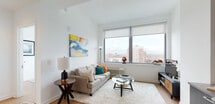 Located at 7 Livingston Ave in the heart of New Brunswick, NJ, this modern residential unit offers a thoughtfully designed single-floor layout with contemporary finishes throughout. Light hardwood flooring flows seamlessly across the living, kitchen, and bedroom areas, creating a cohesive and stylish aesthetic.

The living area is bright and airy, anchored by large floor-to-ceiling windows that frame sweeping views of the New Brunswick skyline, flooding the space with natural light. The open-concept kitchen is outfitted with dark espresso cabinetry, stainless steel appliances including a refrigerator, dishwasher, microwave, and range, complemented by a sleek tile backsplash and ample counter space. A built-in wine rack adds a refined touch near the entryway.

The bedroom is a standout feature, boasting corner floor-to-ceiling windows on two walls that deliver expansive, elevated city views and exceptional natural light. The bathroom is clean and functional, featuring large-format neutral floor tiles, a dark wood vanity with a white countertop, a full-size mirror, and a tub-shower combination with tile surround.

The unit's elevated position within the building, contemporary design, and abundant natural light make it a well-rounded urban living space in one of New Jersey's most dynamic cities.
