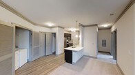 This single-floor residence at 1701 Atria Ct in Raleigh, NC, offers a well-designed open-concept layout that seamlessly connects the kitchen and living areas. The kitchen is a standout feature, centered around a peninsula-style island with a white countertop and undermount sink, complemented by dark cabinetry and stainless steel appliances, including a refrigerator, microwave, and range. Stylish pendant lighting hangs above the island, adding a contemporary touch. The backsplash introduces a subtle textural element that enhances the kitchen's modern aesthetic. Crown molding runs along the upper walls, lending a refined architectural detail throughout the space. The flooring transitions from wood-style planks in the kitchen and entryway areas to plush carpeting in the living space, creating distinct yet cohesive zones. Multiple doors visible along one wall suggest ample storage or closet space. Neutral gray tones on the walls and doors provide a versatile, move-in-ready palette. The layout feels open and functional, making efficient use of the available space while maintaining a clean, modern ambiance throughout.