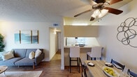 This single-story residence offers a thoughtfully arranged open-concept floor plan that connects the living, dining, and kitchen areas into one cohesive space. Hardwood-style flooring flows throughout the main living areas, complemented by neutral-toned walls and textured ceilings that lend a warm, inviting atmosphere.

The living room is a comfortable gathering space featuring a ceiling fan with integrated lighting, a sliding glass door fitted with vertical blinds that allows natural light to fill the room, and wall-mounted shelving for display. A dedicated dining area sits adjacent to the kitchen, which is defined by a peninsula breakfast bar with seating for two. The kitchen itself is efficiently designed, featuring dark cabinetry, a double stainless steel sink, white appliances including a range and refrigerator, and overhead lighting.

The bedroom is a well-proportioned retreat with hardwood-style flooring, a ceiling fan, and a window dressed with sheer curtains that allow soft, diffused natural light. The bathroom includes a vanity with a large mirror, built-in shelving, and a shower-tub combination with a curtain. Throughout, the space maintains a clean, functional aesthetic suited for comfortable everyday living.