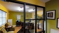 This single-floor space offers a functional and well-appointed layout distinguished by its use of glass partition walls with dark metal frames, creating a sense of openness while defining distinct work areas. The interior is characterized by bold olive-green painted walls that lend a warm, energetic ambiance throughout. Overhead fluorescent lighting ensures consistent, bright illumination across the workspace.

The office areas are furnished with dark wood desks and chairs, providing a professional yet approachable aesthetic. A large decorative indoor tree in a tall floor vase adds a natural element and visual interest to one section of the space. Framed artwork on the walls contributes to an inviting atmosphere.

The flooring transitions between hardwood and area rugs, adding texture and delineating different zones within the space. Office equipment, including a printer or copier, is visible on shared surfaces, indicating a practical, working environment. Additional amenities such as a floor lamp and small appliances suggest the space is fully equipped for day-to-day professional use. Overall, the layout balances productivity with a comfortable, design-conscious interior.