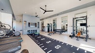 Located at 3 Whisperwood Way in Candler, NC, this single-floor fitness facility offers a comprehensive workout environment thoughtfully designed for a wide range of training needs. The main gym area is expansive and bathed in natural light, thanks to large floor-to-ceiling glass entry doors, transom windows, and multiple wall-mounted windows that provide views of the surrounding outdoor landscape. Recessed lighting and dual ceiling fans maintain a bright, comfortable atmosphere throughout.

The primary workout space features a long turf sprint track runner laid across the polished concrete floor, flanked by a full dumbbell rack with benches, a power rack, and an array of cardio equipment including treadmills, an elliptical, a stair climber, and a stationary bike. Wall-mounted flat-screen televisions are positioned throughout for entertainment during workouts. A built-in storage unit with cabinetry in a muted blue-gray finish provides organized storage for accessories and towels.

A dedicated bonus studio room is enclosed by sleek black-framed glass partition walls and doors, creating a distinct space ideal for barre, stretching, or group fitness. This room features floor-to-ceiling mirrors, a wall-mounted display, ceiling fan, and freestanding barre rails. The overall design is modern and clean, with a neutral color palette, high ceilings, and an open layout that promotes both functionality and an inviting workout environment.
