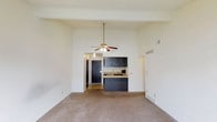 This single-floor residential space showcases an open-concept design that maximizes the sense of roominess and functionality. The living area is defined by impressively high ceilings that lend an airy, expansive feel to the room, complemented by a classic ceiling fan with integrated lighting that provides both comfort and illumination. The flooring transitions from soft, neutral-toned carpet in the main living area to hard flooring near the entryway, creating a natural delineation between spaces. The kitchen is positioned at the heart of the unit, featuring dark wood cabinetry that offers ample storage and a strong visual contrast against the light-toned walls. A peninsula-style breakfast bar serves as both a functional workspace and a casual dining area, seamlessly connecting the kitchen to the living space. A built-in sink is visible within the kitchen, along with upper cabinetry that maximizes vertical storage. The entryway, framed by a dark solid-panel front door, is visible from the main living area, reinforcing the open sightlines throughout. Neutral wall tones and recessed ventilation contribute to a clean, understated aesthetic throughout the space.