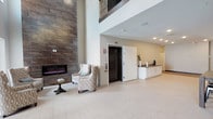 This thoughtfully designed communal space makes a strong first impression in its expansive lobby, where soaring double-height ceilings draw the eye upward to an overlooking upper-level balcony with wrought iron railings. A striking floor-to-ceiling stone tile wall anchors the room, framing a sleek linear electric fireplace that serves as a dramatic focal point. Comfortable lounge seating and a side table invite residents or guests to relax in this welcoming area. A centrally located elevator provides convenient vertical access, while a white cabinetry wet bar or service counter nearby adds a practical amenity to the common area. Light tile flooring throughout the lobby enhances the open, airy feel of the space. A carpeted corridor with geometric patterned flooring connects to additional rooms, including a private conference or meeting room enclosed by dark-framed glass walls and doors. This room features a marble-topped table, upholstered blue chairs, textured striped wallcovering, and a curated gallery wall, all illuminated by a statement drum pendant light. Beyond the corridor, a glass-fronted dining area is visible, furnished with multiple tables and chairs and filled with natural light, completing this versatile and elegantly appointed facility.