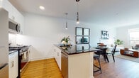 This thoughtfully designed single-floor residence offers a clean, modern aesthetic throughout, with warm hardwood flooring and crisp white walls creating a bright, cohesive atmosphere. The open-concept kitchen is outfitted with sleek white cabinetry, stainless steel appliances including a French door refrigerator, gas range, and microwave, complemented by a dark-toned peninsula countertop. Stylish pendant lights hang above the peninsula, which doubles as a breakfast bar with seating. The dining area flows naturally from the kitchen, offering space for a glass-top dining table and anchored by a striking grid of square wall mirrors that add depth to the room. The living room is inviting and light-filled, featuring a large window with views of the surrounding neighborhood and warm hardwood flooring underfoot. The bedroom is serene and well-proportioned, with natural light filtering through a sizable window, ample closet storage behind double doors, and hardwood floors. The bathroom presents a refined finish with large-format tile flooring, a wood-toned vanity paired with an illuminated mirror, and a wall-mounted storage cabinet, creating a calm and functional space.