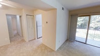 This single-floor space features a straightforward, functional layout with clearly defined living and kitchen zones. The main living area is finished with wall-to-wall carpet in a neutral tone, creating a warm and cohesive feel throughout. Smooth, cream-colored walls and a standard ceiling height contribute to a clean, understated aesthetic.

A partial dividing wall separates the living area from the kitchen corridor, which transitions into tile flooring — a practical distinction between the two spaces. The kitchen is equipped with a white refrigerator and benefits from its own dedicated area, offering a sense of separation without being fully enclosed.

A bi-fold closet door provides convenient built-in storage accessible from the main living space. On the opposite side of the room, a sliding glass door fitted with vertical blinds offers access to an outdoor area and allows natural light to filter into the interior. A ceiling vent indicates a central HVAC system. Overall, the space presents a simple, livable environment with practical finishes and a neutral palette suitable for a variety of furnishing styles.
