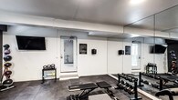 Located in the heart of Hollywood at 1757 N Orange Dr, Los Angeles, this single-floor fitness center is a purpose-built workout facility designed to support a well-rounded exercise routine. The space is divided into two distinct zones, each tailored to different types of training. The first area features clean white walls, rubber flooring, and wall-mounted mirrors that enhance the sense of space. It is outfitted with a rack of dumbbells in varying weights, a set of kettlebells, a flat adjustable workout bench, and a wall-mounted flat-screen television for entertainment during training sessions. A hand sanitizer dispenser and posted signage indicate an attention to cleanliness and facility guidelines. The second zone takes on a darker, more focused aesthetic with charcoal-gray walls and high horizontal windows that allow natural light to filter in. This area houses a NordicTrack Commercial treadmill, a stationary bike, a medicine ball rack with multiple weighted balls, and an additional wall-mounted television. The rubber mat flooring throughout provides both durability and safety. Together, the two areas create a cohesive, functional fitness environment suited for cardio, strength, and conditioning workouts.