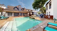 This outdoor amenity area centers around a generously sized swimming pool with clear turquoise water, surrounded by a spacious concrete sundeck designed for relaxation and recreation. Depth markers are clearly visible along the pool's edges, and a raised spa or wading feature sits adjacent to the main pool, connected by a series of stepping stone-style entries that add visual interest to the layout.

The sundeck is furnished with teal-colored lounge chairs and small side tables, providing comfortable spots for sunbathing. A wooden pergola structure anchors one side of the pool area, offering shaded outdoor seating beneath its open lattice roof. Flanking the pergola is a building with large French-style windows and doors that open directly onto a raised wooden deck, creating a seamless indoor-outdoor connection.

The surrounding multi-story residential buildings feature a mix of bold navy blue and neutral white exteriors with pitched, shingled rooflines, multiple chimneys, and exterior staircases. Mature trees are scattered throughout the courtyard, providing natural shade and greenery. The entire pool area is enclosed by black metal fencing, creating a defined and secure outdoor retreat with a resort-like ambiance.
