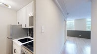 This studio in Memphis, TN (38104) offers a thoughtfully designed single-floor layout comprising a living room, kitchen, bathroom, and hallway. The living area is bright and open, featuring light wood laminate flooring, a textured ceiling, and a sleek black ceiling fan. A row of wide windows with horizontal blinds floods the space with natural light, while a distinctive dark wood shiplap accent wall running beneath the windows adds a modern, layered design touch. Sliding closet doors provide practical storage within the living space. The kitchen is efficiently arranged with white shaker cabinetry, a stainless steel Frigidaire refrigerator, a stainless gas range, a built-in microwave, a black pull-down faucet, and a dark hexagonal tile backsplash that adds visual interest. Exposed rail-style ceiling tracks run through the hallway connecting the kitchen to the main living area. The bathroom is a standout feature, showcasing a soaking tub with marble-effect wall surround panels and matte black fixtures, paired with a slatted wood vanity base for a refined, contemporary aesthetic. Throughout the space, the consistent use of light tones, black hardware accents, and warm wood flooring creates a cohesive and modern atmosphere.
