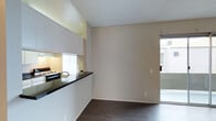 This single-floor space offers a clean, modern aesthetic characterized by an open-concept design that seamlessly connects the kitchen and living areas. The kitchen is well-appointed with sleek white cabinetry extending to the ceiling, providing generous storage. A dark granite countertop serves as a breakfast bar, creating a natural divide between the kitchen and the adjoining living space. Under-cabinet lighting adds a warm, functional glow to the cooking area, which is equipped with a gas range. A mirrored backsplash enhances the sense of depth and brightness within the kitchen. The living area features neutral-toned walls and dark hardwood-style flooring, which grounds the space with a rich, contemporary contrast. A sliding glass door at the far end of the living area floods the room with natural light and provides direct access to an outdoor area, offering a connection to the exterior. The textured ceiling adds subtle architectural character throughout. The overall ambiance is modern and understated, with a cohesive palette of whites, grays, and dark tones that lend the space a polished, move-in-ready quality.