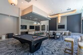 Located at 1240 La Avenida St in Mountain View, CA, this upscale amenity lounge is a thoughtfully designed communal space spanning a generous single-floor layout. The interior immediately impresses with soaring double-height ceilings and an abundance of natural light streaming through expansive floor-to-ceiling windows that frame views of an outdoor pool and surrounding multi-story building.

A dramatic floor-to-ceiling marble-clad accent wall anchors the central gathering area, where a large round dining table with upholstered chairs invites group meals and social events. Adjacent to this is a billiards area with teal upholstered booth seating, adding a vibrant, playful contrast to the neutral palette.

The fully equipped kitchen is a standout feature, showcasing a substantial waterfall-edge quartz island with bar seating, dark wood cabinetry with gold hardware, stainless steel appliances including a French door refrigerator, and a white brick-tile backsplash. Dual flat-screen televisions are wall-mounted above the counter for entertainment.

The lounge area features plush sofas arranged over a bold geometric-patterned area rug, complemented by stylish pendant lighting in white and gold tones. Porcelain tile flooring throughout ties the open-plan space together. Smart home control panels are discretely integrated into the walls, reflecting the building's modern technological infrastructure.