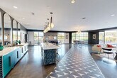 Located at 726 Fulton St in Chattanooga, TN, this single-floor community space offers a generously sized, open-plan interior that seamlessly blends a dining room and living room into one cohesive environment. The space is defined by its contemporary design language, featuring polished concrete floors punctuated by a bold geometric patterned tile runner that anchors the central gathering area. A striking long white island with a decorative scalloped base serves as a functional centerpiece, complemented by pendant lighting with leather-wrapped fixtures that add warmth and character overhead. A separate wet bar area features vibrant teal cabinetry, white quartz countertops, and a series of arched mirrors that visually expand the space. The dining zone is furnished with sleek bar-height tables and modern stools beneath globe chandelier lighting, while large floor-to-ceiling windows and glass curtain walls flood the interior with natural light and provide views of the surrounding streetscape and outdoor pool area. A living lounge area showcases a textured geometric wood-paneled accent wall with a wall-mounted screen and a linear fireplace, adding a sophisticated focal point. Folding glass doors connect seamlessly to an outdoor pool deck, making this a highly versatile and inviting communal destination.